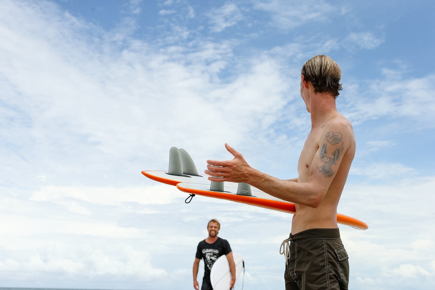 Troy Elmore holding an orange surfboard overhead against a bright sky with its quad fins fitted, another surfer walking up the beach behind him