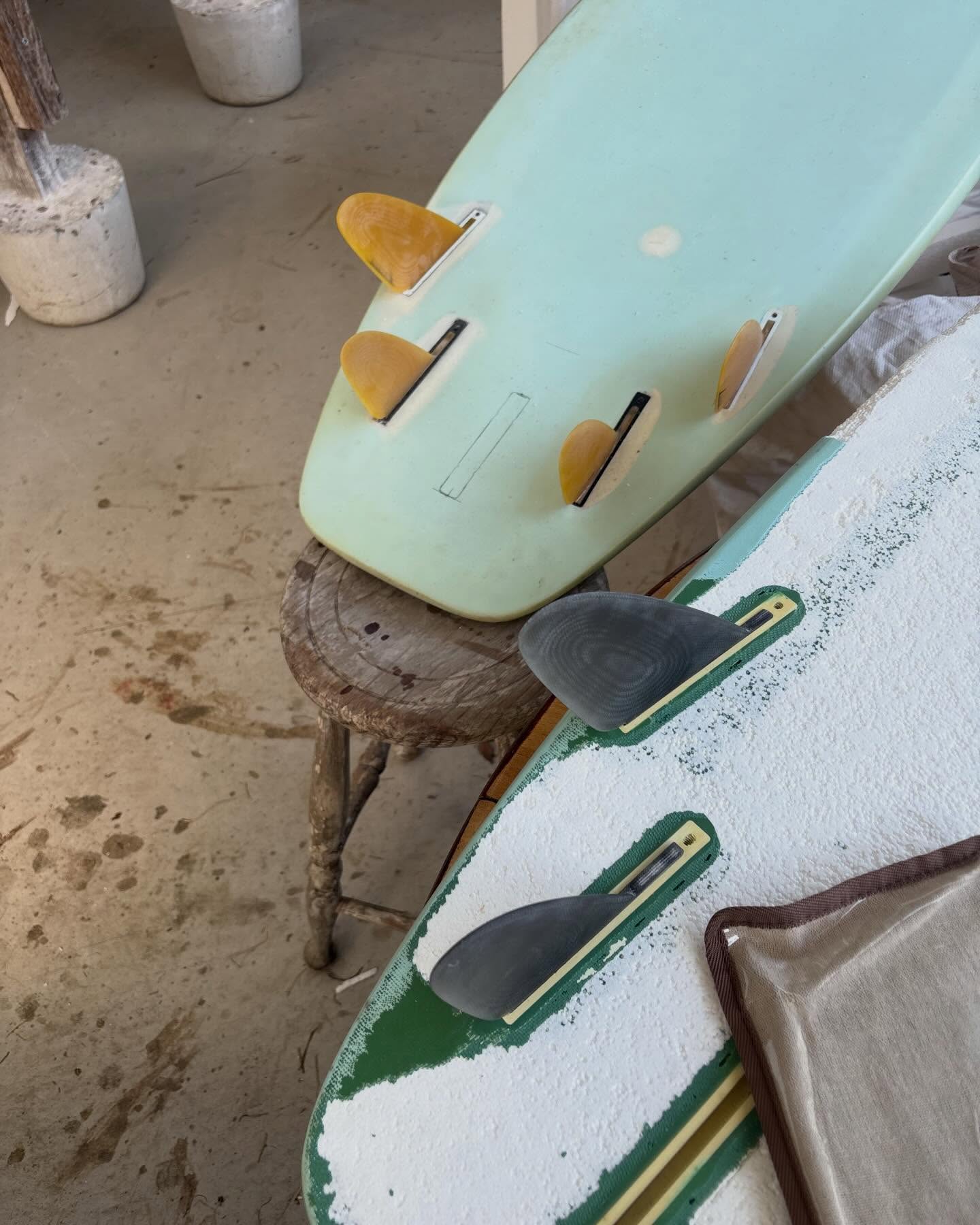 The tail of a mint green surfboard in a shaping bay with four amber fins fitted β two larger fronts and two smaller rears β and the centre fin box left empty