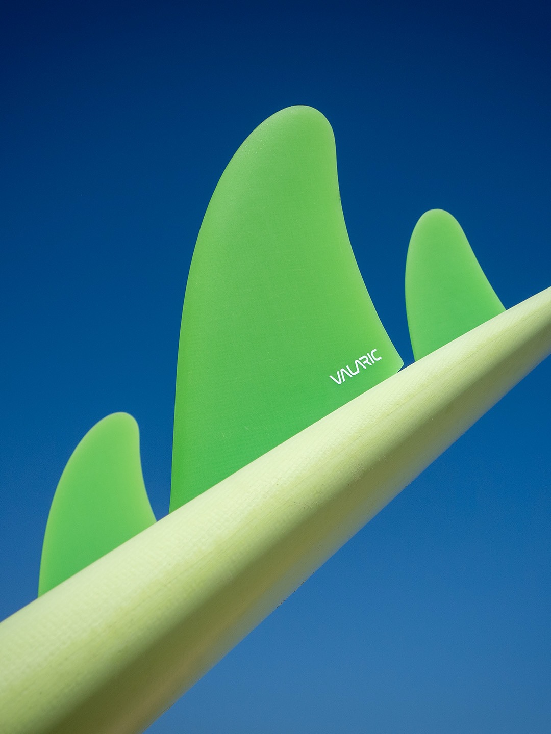A Valaric quad set on the rail of a pale surfboard against a deep blue sky β a tall front fin beside a much smaller rear fin, showing how unevenly a quad's area is distributed